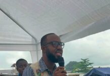 CEEPARD Supports 2,500 Vulnerable Children with Learning Materials in Waterloo Man wearing a beige utility vest and glasses speaks into a handheld microphone under a white canopy at an outdoor event, gesturing with his other hand.
