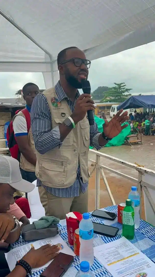 Man wearing a beige utility vest and glasses speaks into a handheld microphone under a white canopy at an outdoor event, gesturing with his other hand.