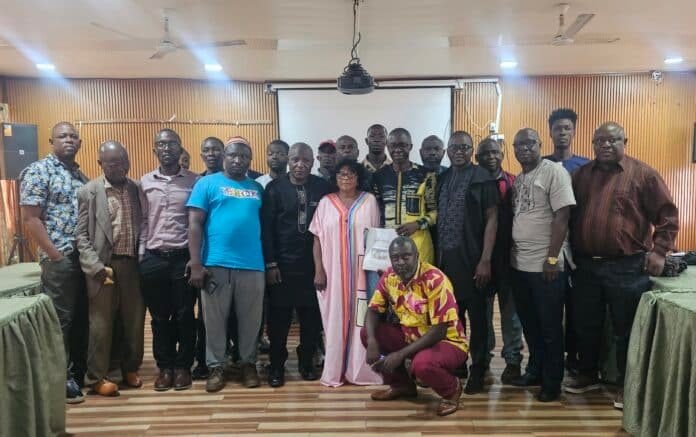 Group of around 20 adults posing for a photo in a wood-paneled room, some in traditional attire, others in casual or business clothing. A projector screen hangs in the back. Two people kneel at the front while everyone stands close together, smiling for the camera.