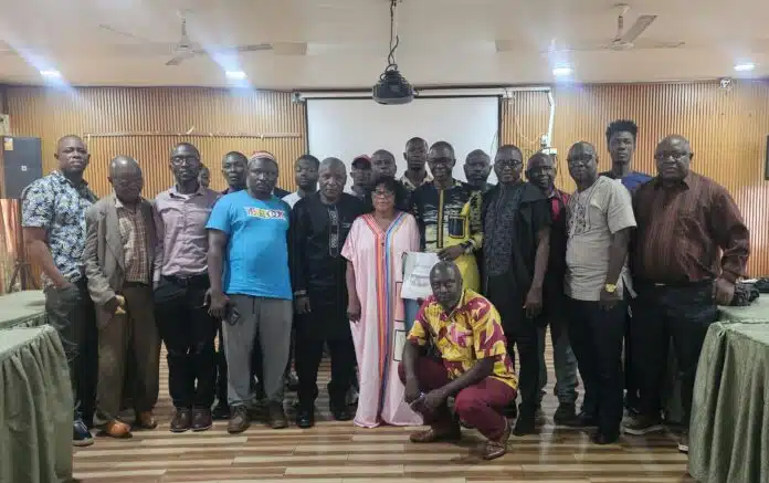 Group of around 20 adults posing for a photo in a wood-paneled room, some in traditional attire, others in casual or business clothing. A projector screen hangs in the back. Two people kneel at the front while everyone stands close together, smiling for the camera.