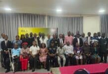 Amnesty Seeks Shift to Notification System for Public Processions Group of diverse men and women posing for a formal photo in a conference room, with a yellow Amnesty International banner in the background.