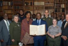 President Bio Invokes Amistad Legacy to Renew Sierra Leone’s Call for Reparatory Justice Group of diverse officials and professionals standing in a wood-paneled library, a man in a blue suit holding an open book toward the camera.