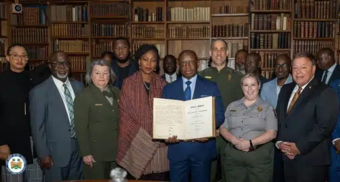 Group of diverse officials and professionals standing in a wood-paneled library, a man in a blue suit holding an open book toward the camera.