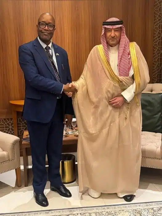 Two men in formal attire shake hands in a room with wood panels and beige furniture, signaling a meeting or agreement.