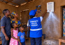 Sierra Leone Deploys 101,862 Spatial Emanators to Strengthen Malaria Prevention Community health worker in a blue vest demonstrates a medical device mounted on a mud-walled wall to a family of four in a rural home.