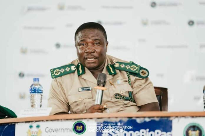Sierra Leone government official in uniform speaks into a microphone at a press conference with a bannered table in front.