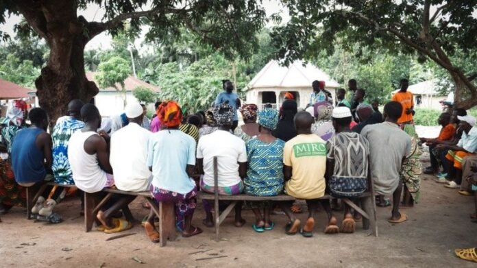 Community meeting under a large tree with people seated on benches listening to a speaker.