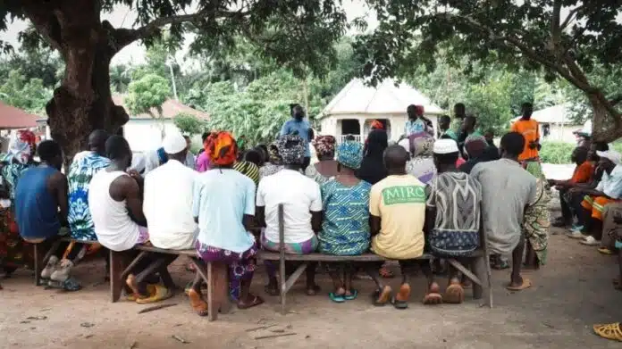 Community meeting under a large tree with people seated on benches listening to a speaker.