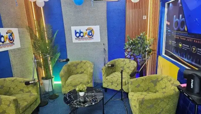 Radio studio interview set with four green patterned chairs around a black marble coffee table, microphones on stands, plants, and branding on blue/gray walls.