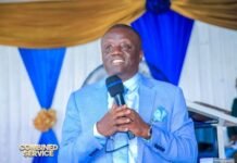 Pastor Francis Mambu Unshaken by Social Media Attacks, Stands Firm on God’s Word Man in a light-blue suit speaks into a wireless microphone at a formal event; decorative blue and gold backdrop with a Combined Service logo in the corner.