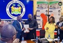 New Finance Minister Karefa Kargbo Pledges Accountability, Reforms and Delivery Two men exchange a document at a Ministry of Finance meeting while colleagues applaud; a government emblem is on a screen behind them.