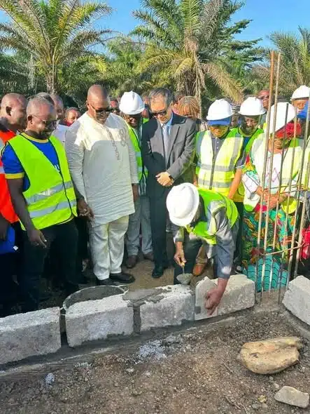 Group of construction workers and officials in safety vests and hard hats observe as a crew member places a concrete block at a construction site, with palm trees in the background.