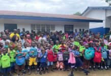 Dr. Walton Gilpin’s GEED Foundation Empowers 300 Children with School Materials A large group of schoolchildren with colorful backpacks standing outside a classroom building.