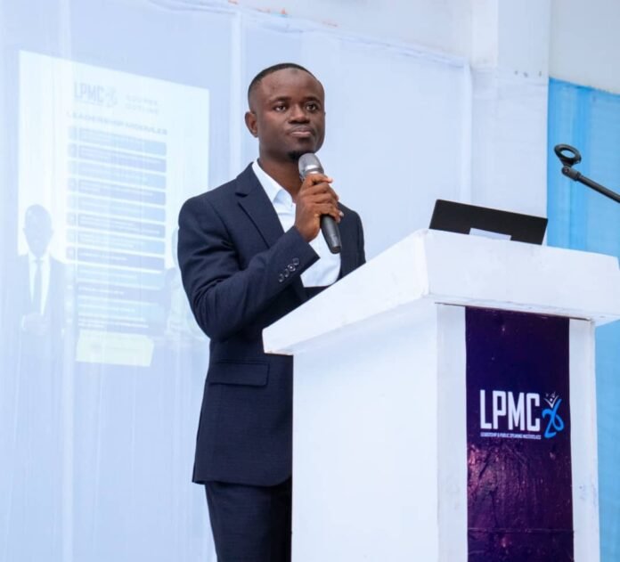 Man in a dark suit speaking into a handheld microphone at a white podium with a laptop, LPMC logo on the front panel.
