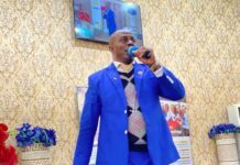 From the Pulpit to Public Controversy: Why Apostle Lelleh’s Right to Preach Must Be Respected Man in a bright blue suit speaks into a microphone on a stage with ornate wallpaper and flower arrangements nearby.