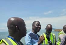 Labour Congress Declares Port Congestion an Emergency, Backs Speedy Completion of Gento Port Group of men wearing high-visibility vests stand near a rocky coastline; one man speaks into a handheld microphone for a news interview on a sunny day.