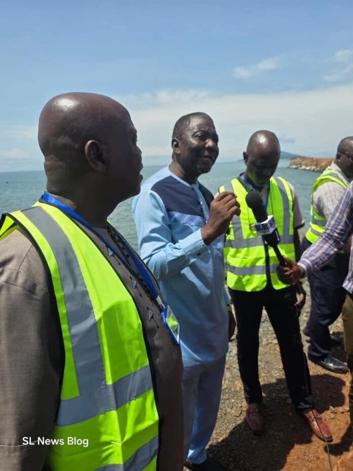 Group of men wearing high-visibility vests stand near a rocky coastline; one man speaks into a handheld microphone for a news interview on a sunny day.