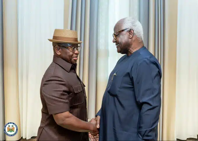 Two older men shake hands and smile in a room with cream curtains; one wears a brown shirt and straw hat, the other in a dark blue traditional outfit.