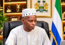 Alhaji Amadu Juldeh Sowe Declares for Tabital Pulaaku International Presidency Public official in traditional white attire sits at a desk, with a flag and crest behind him in an office setting.