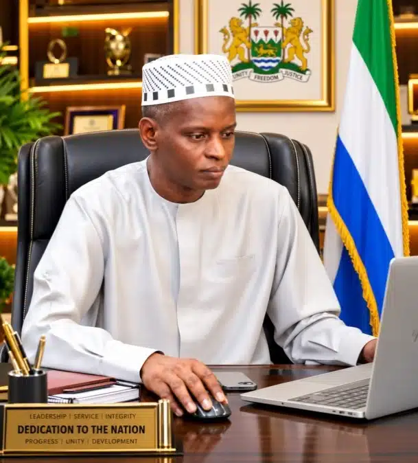 Public official in traditional white attire sits at a desk, with a flag and crest behind him in an office setting.