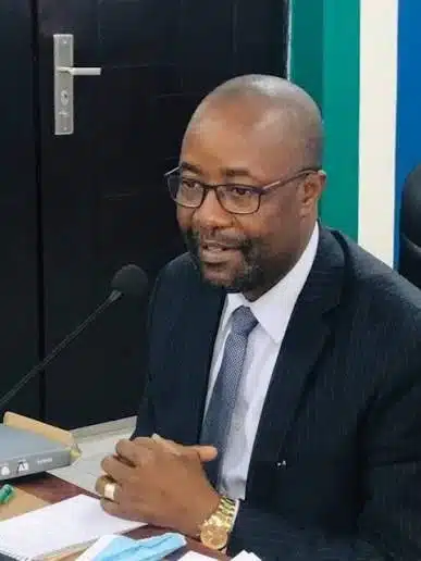 Man in a dark suit and tie, wearing glasses, speaking into a microphone at a desk during a formal meeting.
