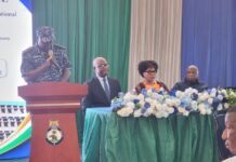 SLP Marks UN International Day of Police Cooperation with Focus on Trust, Accountability Military officer in uniform speaks at a podium as a panel of three dignitaries sits behind a flowered green-table on a stage.
