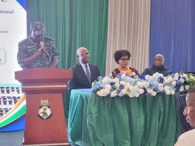 Military officer in uniform speaks at a podium as a panel of three dignitaries sits behind a flowered green-table on a stage.