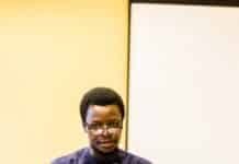 SLAJ Pushes for Stand-Alone Constitutional Chapter on Free Speech, Press Freedom Man wearing glasses and a navy shirt speaks into a blue microphone at a wooden lectern, with panelists seated behind him at a long table.