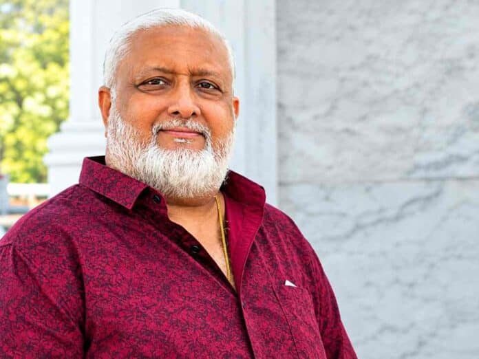 Smiling older man with a white beard in a maroon patterned shirt, standing outdoors near a marble wall.