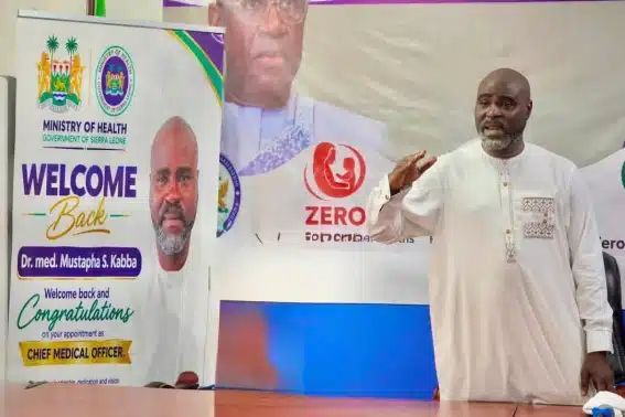 Man in white traditional attire stands at a conference table, speaking or gesturing; behind him are banners from the Ministry of Health welcoming back the Chief Medical Officer, with logos and the words 'WELCOME Back' and 'Chief Medical Officer' visible.
