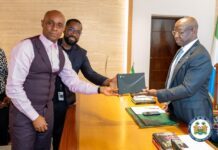 President Bio Receives Flot Card No. 001 Ahead of Official Launch Two men exchange a dark folder across a wooden desk in a formal office setting, with one man in a pink shirt and vest and the other in a suit observing nearby helpers.