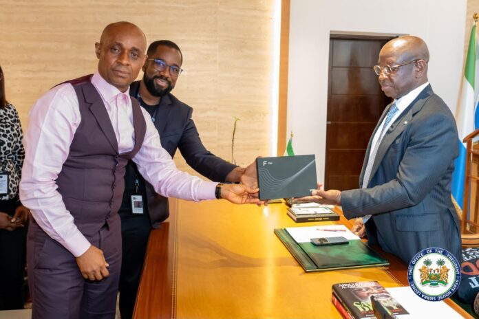 Two men exchange a dark folder across a wooden desk in a formal office setting, with one man in a pink shirt and vest and the other in a suit observing nearby helpers.
