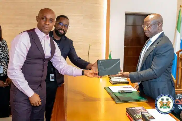 Two men exchange a dark folder across a wooden desk in a formal office setting, with one man in a pink shirt and vest and the other in a suit observing nearby helpers.