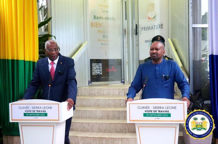 Two officials stand at podiums giving a joint press briefing during a Guinea–Sierra Leone work visit, flags and steps in the background, and a government seal visible to the right.