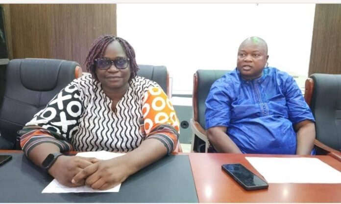 Two adults sit at a conference table in a meeting room, the woman in a patterned blouse and glasses and the man in a blue traditional outfit.