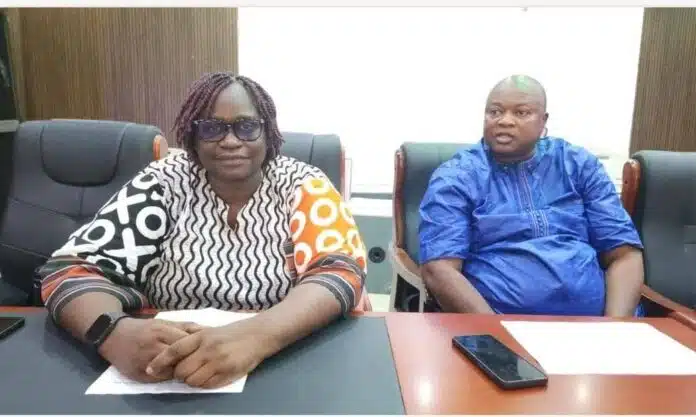 Two adults sit at a conference table in a meeting room, the woman in a patterned blouse and glasses and the man in a blue traditional outfit.