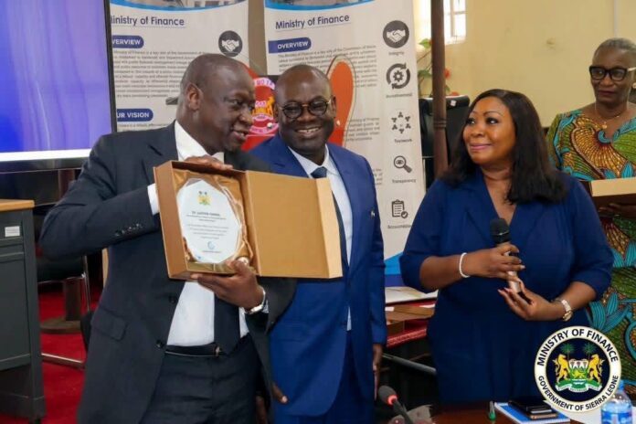 Three professionals at a government awards event; man on the left presents an award in a wooden box to a smiling colleague, while a woman speaks into a microphone in the foreground.