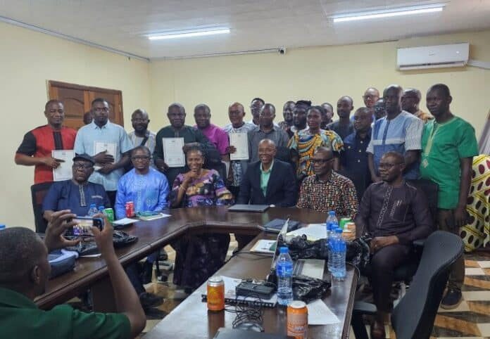Group of about 24 people in a conference room, some holding documents, seated around a U-shaped table with water bottles on it.