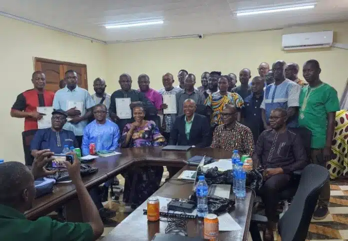 Group of about 24 people in a conference room, some holding documents, seated around a U-shaped table with water bottles on it.