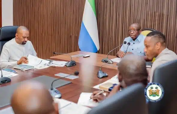 A formal government meeting with officials seated around a wooden conference table, microphones and documents in front of them, and a national flag in the background along with an official seal.