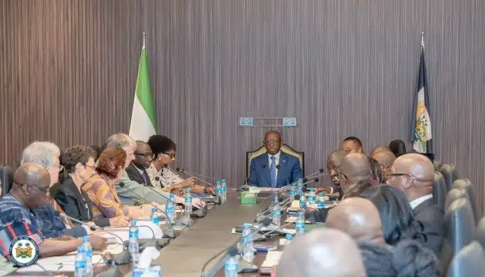 Formal meeting with a man in a blue suit at the head of a long conference table, surrounded by delegates, flags behind him.