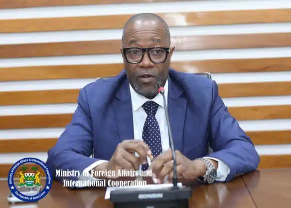 Male government official in a blue suit and glasses speaks into a microphone at a desk during a press briefing for the Ministry of Foreign Affairs and International Cooperation (official seal visible).