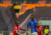From KT Field to The International Stage: Abubakarr Silla a Rising Star from Kingtom’s Journey Two young soccer players in red and blue kits chase a ball on a green pitch during a match, stadium stands in the background.