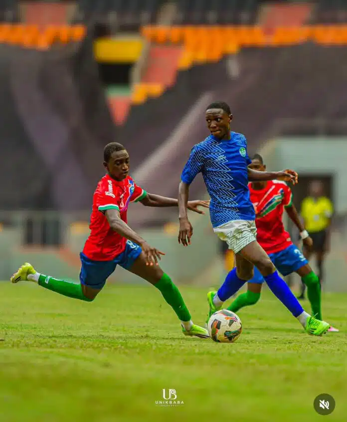 Two young soccer players in red and blue kits chase a ball on a green pitch during a match, stadium stands in the background.