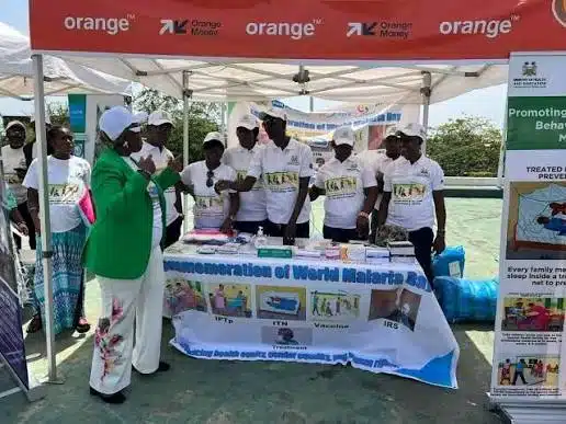 Group of volunteers at an outdoor World Malaria Day information booth under an orange banner.