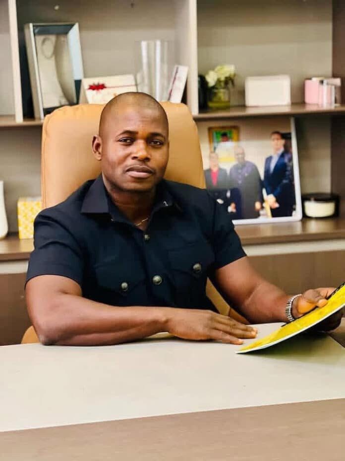 Man seated at a desk in an office, looking at the camera while holding a yellow document or folder in his right hand.