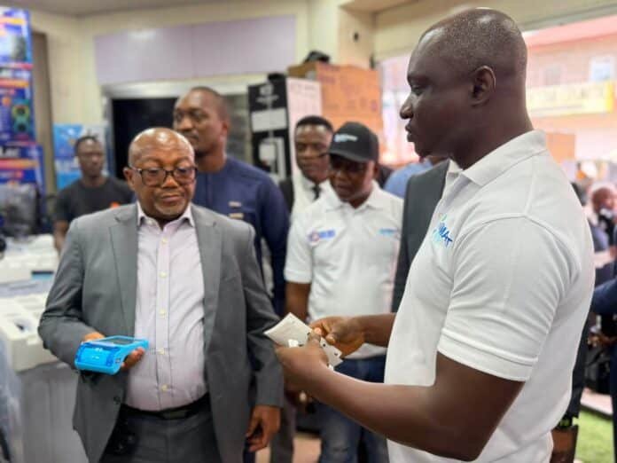 Man in a gray suit hands a blue package to someone in a store, while a group watches in the background.