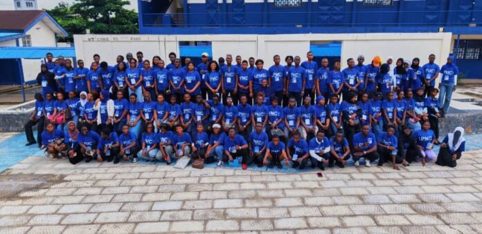 Large group of people in matching blue shirts posing for a group photo outdoors, with a blue and white building in the background.
