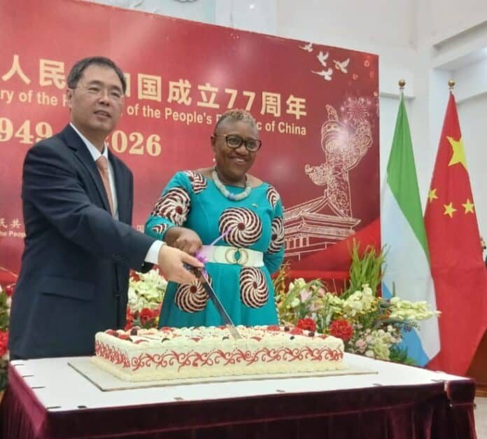 Man and woman cut a large cake at a 77th anniversary celebration for the People's Republic of China, with Chinese flags in the background.