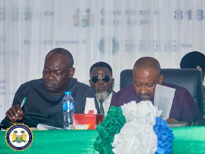 Two men in a formal meeting sit at a green-draped table; one writes with a pen, the other reads a document, with an official seal visible on the left.